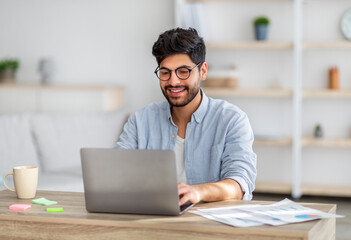 Portrait of happy arab freelancer man sitting at desk with laptop computer at home office, looking and smiling at screen - obrazy, fototapety, plakaty