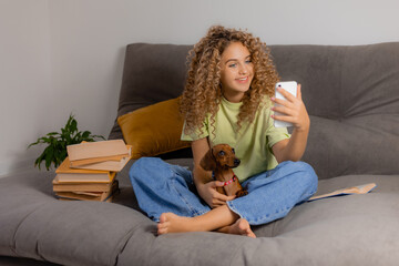 blonde girl with blue eyes and curly hair takes a selfie with a dachshund dog sitting on a gray sofa. young woman takes photos on her smartphone. space for text. High quality photo