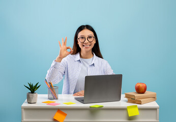 Happy asian female student in eyeglasses showing okay sign gesture sitting at desk and using laptop over blue background