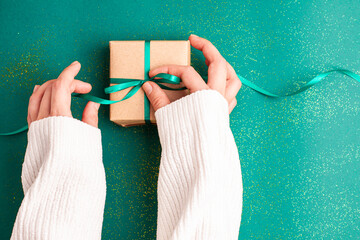Human hands in white sweater tieding brown gift box with a emerald green satin ribbon bow on green background with gold colored glitter. Christmas Holidays concept flat lay