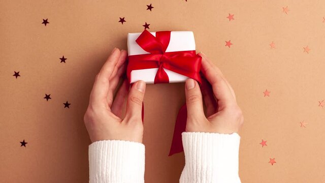Human hands in white sweater puts a white paper gift with a red satin ribbon bow on brown background with red stars shapes. Stop motion animation Christmas Holidays concept flat lay
