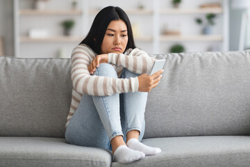 Obraz premium Upset Asian Lady Looking At Smartphone Screen, Sitting On Sofa At Home