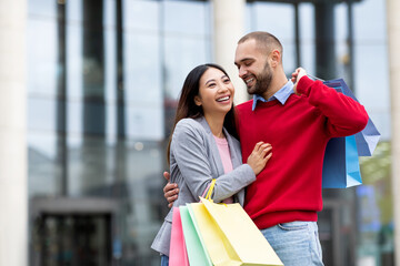 Portrait of joyful interracial couple in casual clothes posing and smiling, holding shopping bags near big supermarket