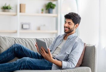 Relaxed arab guy resting on sofa with digital tablet, browsing social networks or shopping online, free space