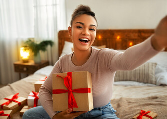 Excited african american woman holding gift box and making selfie, sitting on bed in cozy bedroom...