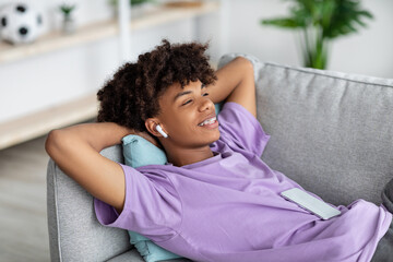 Relaxed black teenager in earphones listening to music or audio book, lying on couch at home