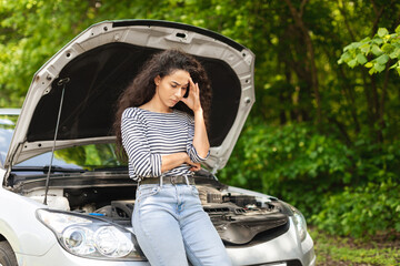 Puzzled woman driver leaning on open car hood