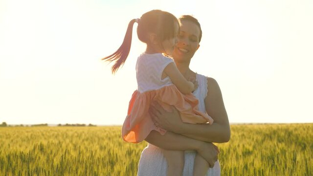 A little daughter in her mother's arms, they walk together in a wheat field, hug and kiss. A happy family, a child and mommy are walking in the park in summer. Farmer woman and kid in the field