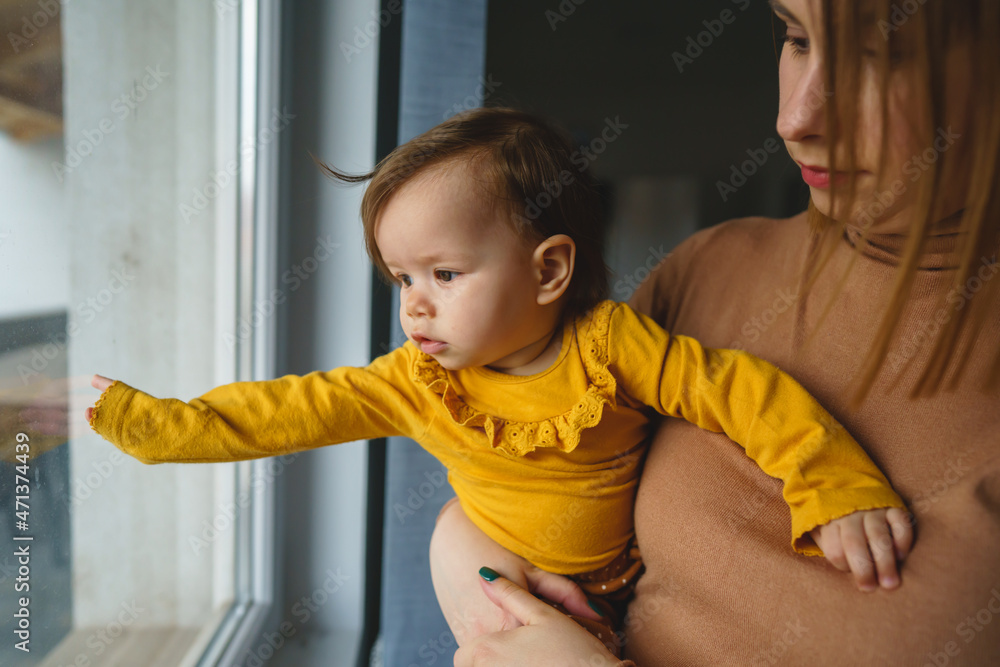 Wall mural One small caucasian baby girl with arms spread in hands of her mother holding her at home in day looking trough the window real people side view copy space family bonding leisure concept