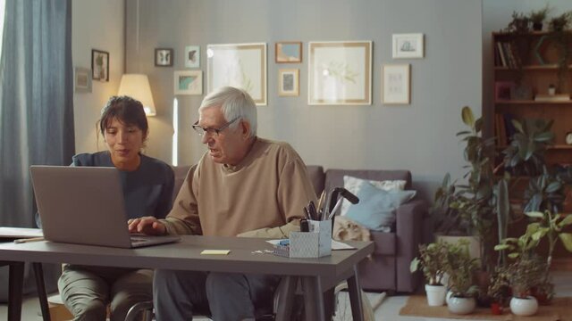 Young woman spending time with senior man at home helping him to learn how to use laptop for Internet surfing and work
