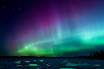 Northern Lights erupt over a Minnesota lake in the dark sky overhead shining a rainbow of Aurora light and colors over the forests and frozen water and ice