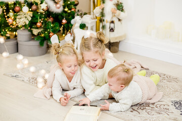 3 blonde girls lie on the floor by the Christmas tree and read a book. cozy evenings with a book by the tree. Girlfriends read a book together. Warm relationship