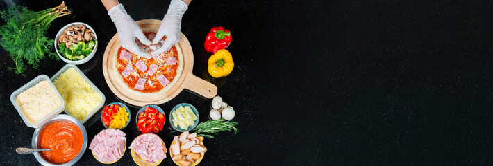 Top view of chef adding ham into the dough of a homemade pizza with tomato sauce while wearing gloves and pizza ingredients, spices on black background. Pizza menu. View from above. Space for text.