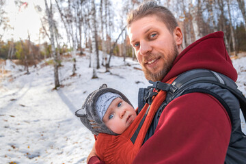 Adorable smiling bearded babywearing father with his baby in sling winter forest walking.