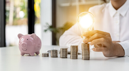 Business Woman holds a glowing light bulb on top of the highest pile of coins, Placing coins in a row from low to high is comparable to saving money to grow more. Money-saving ideas for investing.