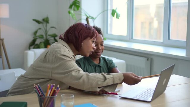 Distant home schooling of junior children. Positive african-american lady shows information on laptop screen to little son at online lesson in living room spbd