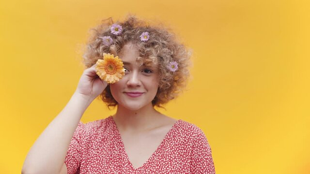 Happy Woman With Daisy Flower In Hand And On Her Blonde Curly Hair Standing On Yellow Orange Background. - Medium Closeup Shot