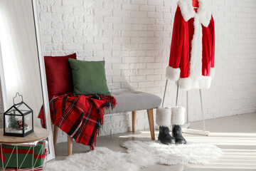 Obraz premium Santa claus costume with boots and christmas decorations in a festive room setting, a santa claus costume with red and white fur trim hangs on a stand, accompanied by black boots with white fur cuffs