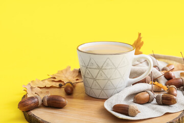 Board with cup of coffee, acorns and autumn leaves on yellow background