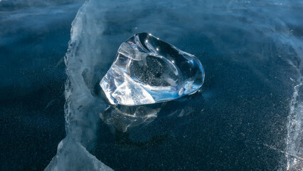 A shiny transparent shard of ice on a blue frozen lake. Deep cracks are visible. Reflection on the surface. The glare of the sun on the edges. Baikal