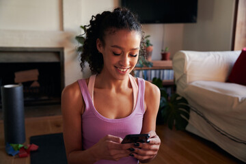 Multi-cultural young adult female smiling and texting while on yoga mat in modern lounge at home