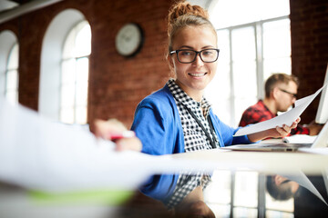 Happy young businesswoman working with papers while sitting by desk on background of colleague