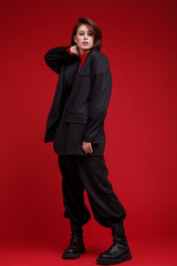 Fashion photo of a beautiful elegant young woman in a pretty oversize black suit, pants, top posing over red background. Bob haircut. Studio Shot. Rebellious free street style. Rebel and classic