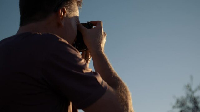 Medium handheld shot of a tourist photographer raising a camera and taking a photo and checks it with late afternoon sun on him in Africa.