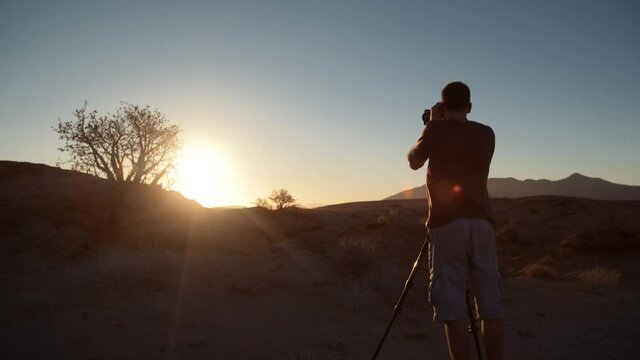 Dramatic wide static handheld shot of  a tourist photographer taking a landscape picture towards the sun on a tripod in a desert in Africa.
