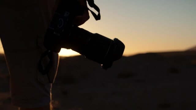 Dramatic tracking shot of a tourist photographer's hand has he carries a camera walking towards the setting sun in a desert in Africa.