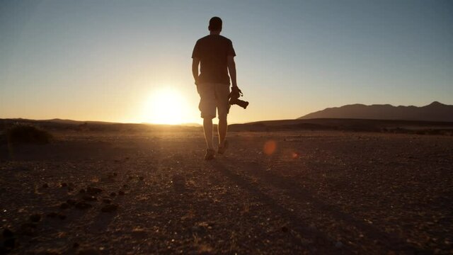 Dramatic wide follow shot of a tourist photographer walking towards the sun in a desert in Africa