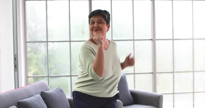 Mature 60s Asian woman dancing with happy and cheerful action during listening to music alone in home living room. Concept of happiness lifestyle and good mental health of senior retirement people.