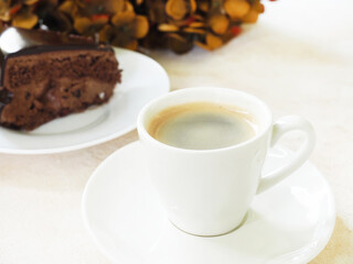 Espresso coffee with foam in a white cup. There are gold brown decorative flowers and a slice of chocolate cake. 