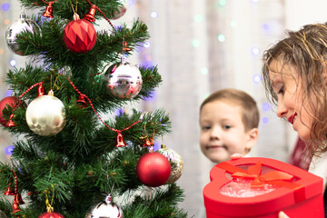 A seven-year-old boy in a smart shirt gives his mother a gift in a box for the New Year against the backdrop of a Christmas tree at home. Selective focus. Portrait