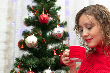 Young beautiful woman in a red sweater with a mug of cocoa and marshmallows at home by the new year tree on new year's eve. Selective focus. Portrait