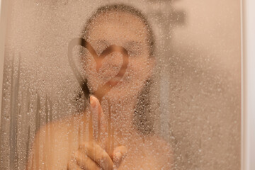 Photo of smiling attractive young woman draws heart on weeping glass shower door, enjoys rest in douche, washing her body, standing behind steam blurred glass with water drops. - obrazy, fototapety, plakaty