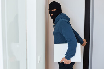 Man robber wearing blue hoodie and black balaclava posing in a robbed house, looking at the camera, holding a stolen laptop, a male person has committed unlawful acts.