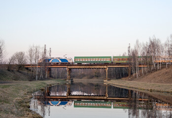 View of a small river in the spring against the background of a passing train with freight cars on a railway bridge. Channel of the Vileyka-Minsk water system for the transfer of water flow