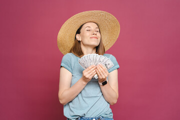 An image of a joyful young lady in a blue summer t shirt and big hat is holding money happily on pink background