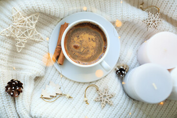 A cup of coffee over Christmas bokeh garlands in the house on the table with a sweater in the background and decorations. Winter mood, festive decoration, magical Christmas.