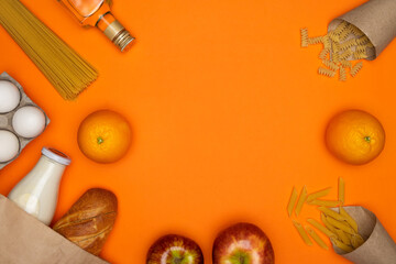 Bread and bottle of milk in paper eco bag. Spaghetti, sunflower oil, eggs and red apple on orange background. Healthy food, delivery, donation concept. Food stock for quarantine. Top view, copyspace.