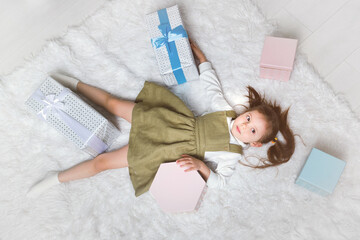Obraz na płótnie Canvas Cute little girl lies on a white carpet with Christmas gifts around