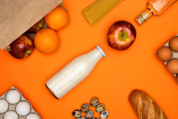 Red apples and oranges in paper eco bag. Spaghetti, bottle of milk, eggs, bread, olive oil on orange background. Healthy food, delivery, donation concept. Food stock for quarantine. Top view, flatly.