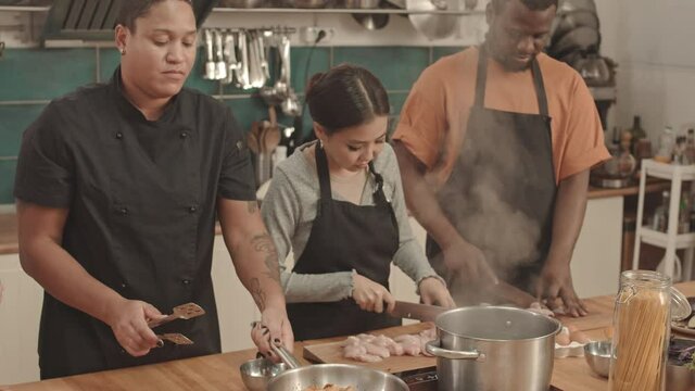 Medium of young female chef roasting chicken in pan on stove, Asian woman and African American man standing by table in large kitchen, cutting fillets, Caucasian sous-chef watching