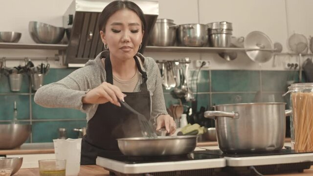 Medium low angle of young Asian woman wearing apron, standing by stove in professional kitchen, adding cream to food in frying pan and mixing