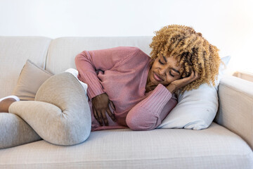 Shot of a young woman experiencing stomach pain on the sofa at home. I need painkiller and a hot water bottle. Young woman in pain lying on couch at home