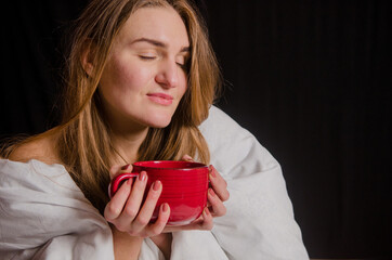 Beautiful young woman with blonde hair, sits in the morning wrapped in a white blanket on black background, drinks tea from a red cup