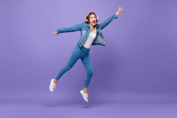 Fototapeta na wymiar Happy Asian woman jumping while listening to music on headphones in isolated purple background