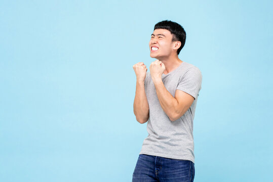Happy ecstatic young Asian man raising his fists doing yes gesture celebrating success in isolated light blue studio background