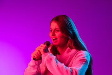 Naklejka premium Young smiling beautiful girl singing with microphone isolated on purple studio background in neon pink filter. Concept of emotions,
